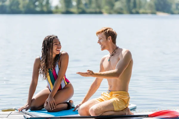 Young and redhead man pointing with hand and talking to cheerful african american woman in colorful swimsuit while sitting together on sup boards on lake — Stock Photo