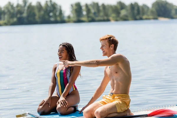 Excitado pelirrojo hombre apuntando con el dedo cerca sorprendido africano americano mujer en traje de baño colorido mientras se sientan juntos en tablas de sup en el río en verano - foto de stock