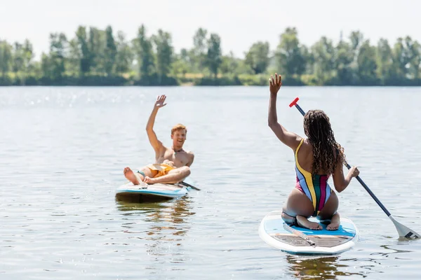 Brunette african american woman in striped swimsuit and overjoyed redhead man waving hands at each other while sailing on sup boards on lake in summer — Stock Photo