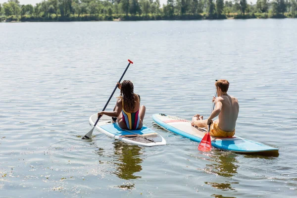 Back view of brunette african american woman in striped swimsuit sailing on sup board near redhead and sportive man on picturesque lake during summer day — Stock Photo