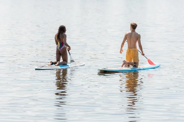 Back view of sportive and redhead man and african american woman in striped swimsuit kneeling on sup boards and sailing on river on summer vacation day — Stock Photo