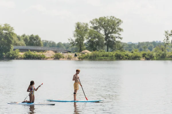 Vista lateral de un joven hombre deportivo y una mujer afroamericana de pie sobre las rodillas y navegando en tablas de sup con paletas en el río con banco verde en verano - foto de stock