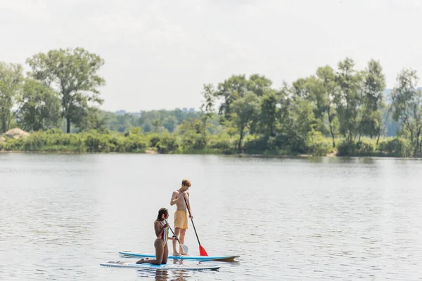 Active multiethnic couple in colorful swimwear sailing on sup boards with paddles while spending summer vacation day on river with green trees on scenic bank — Stock Photo