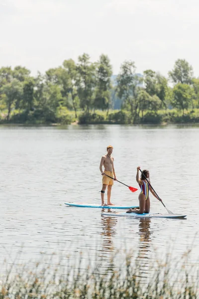 Redhead and sportive man paddleboarding along green riverside near african american woman in colorful swimsuit during summer recreation on summer weekend — Stock Photo