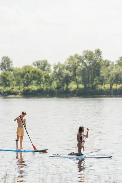 Side view of young redhead man and african american woman in colorful swimsuit spending summer weekend while sailing on sup boards on picturesque river — Stock Photo