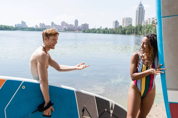 Happy redhead man holding sup board and talking to enchanting african american woman in colorful swimsuit near lake and cityscape on background in summer — Stock Photo