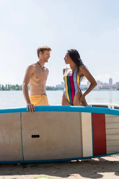 Smiling redhead man looking at brunette african american woman in colorful swimwear standing with hand on hip near sup board with river and cityscape on background — Stock Photo