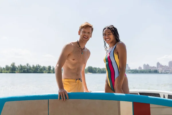 Appealing african american woman in striped swimsuit and young redhead man in yellow swim shorts looking at camera near sup boards with lake and cityscape on background — Stock Photo