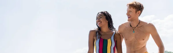 Mujer afroamericana llena de alegría en traje de baño colorido y joven pelirrojo mirando hacia otro lado y sonriendo bajo el cielo azul durante el fin de semana de verano al aire libre, pancarta - foto de stock