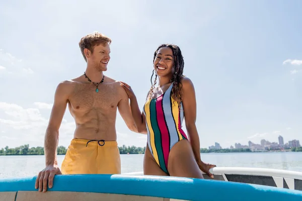 Cheerful and enchanting african american woman in colorful swimsuit looking at camera near young and redhead man and sup boards under blue sky outdoors — Stock Photo