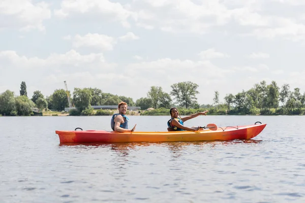 Беззаботная африканская американка в спасательном жилете указывает вперед и улыбается в камеру во время плавания на спортивном байдарке с рыжей подругой во время летних выходных на реке — стоковое фото