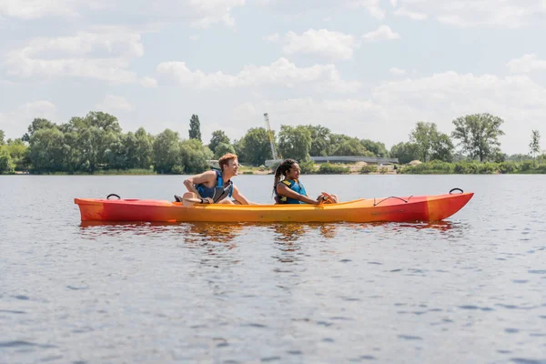 Side view of brunette african american woman and young redhead man in life vests paddling in sportive kayak on lake with green shore under blue sky with clouds — Stock Photo