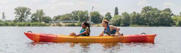Side view of sportive and interracial couple in life vests sailing in kayak during summer water recreation on river with green picturesque bank, banner — Stock Photo