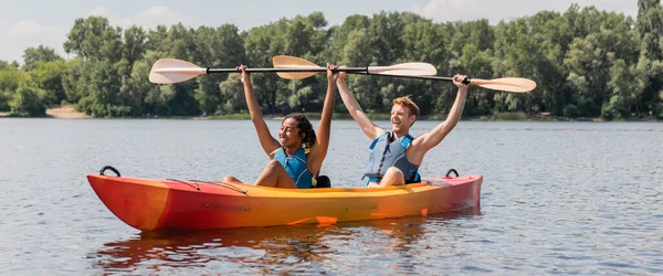 Activa y feliz pareja multiétnica en chalecos salvavidas levantando paletas en las manos mientras se sienta en kayak deportivo en el río con árboles verdes en el fondo en el día de verano, bandera - foto de stock