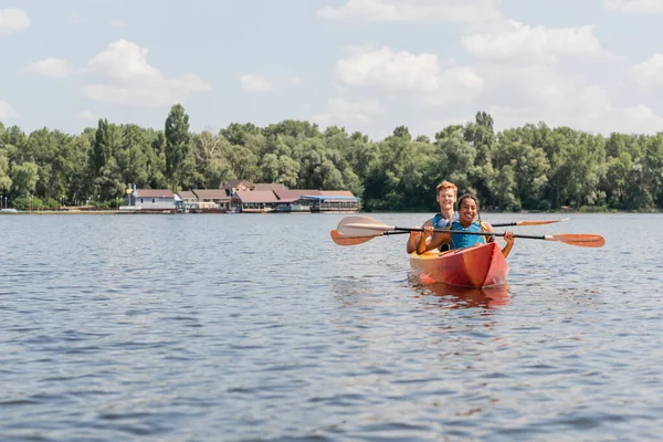 Allegra donna afroamericana e giovane uomo dai capelli rossi in giubbotti di salvataggio remare in kayak sportivo mentre trascorre del tempo sul fiume durante il fine settimana nella giornata estiva — Foto stock