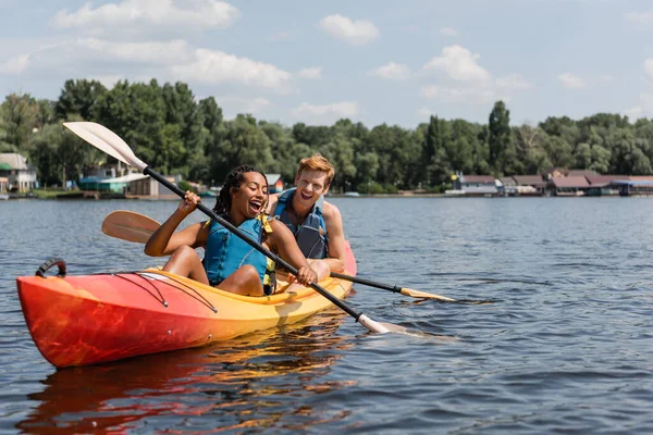 Overjoyed african american woman and young redhead man in life vests spending summer weekend by sailing in kayak with paddles on lake with green scenery shore — Stock Photo