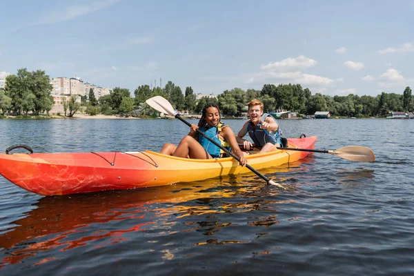 Joven y feliz pelirroja en chaleco salvavidas señalando con el dedo y remando en kayak con la encantadora mujer afroamericana en el lago con la orilla verde en verano - foto de stock
