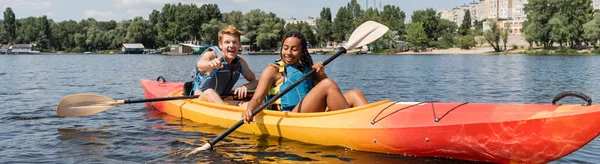 Homme rousse ravi pointant du doigt près de femme afro-américaine en gilet de sauvetage tenant la pagaie tout en naviguant en kayak le long de la rivière verte en été, bannière — Photo de stock
