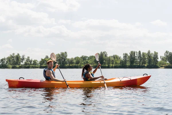 Side view of smiling redhead man and brunette african american woman in life vests spending summer weekend by sailing in kayak on river under cloudy sky in summer — Stock Photo
