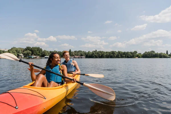 Carefree and charming african american woman and young redhead man in life vests sailing in sportive kayak on calm water surface under blue sky with clouds on summer day — Stock Photo