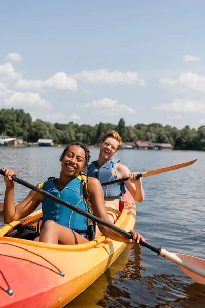 Возбужденная многонациональная пара в спасательных жилетах проводит летние выходные на живописном озере, катаясь на спортивном байдарке под голубым небом с белыми облаками — стоковое фото