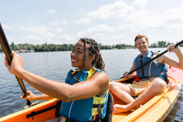 Cheerful african american woman in life vest sailing in kayak while spending time on river with young and redhead man paddleboarding on blurred background — Stock Photo