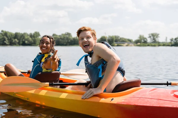 Eccitata rossa uomo con bocca aperta guardando la fotocamera vicino allegra donna africana americana in giubbotto di salvataggio che punta con il dito durante la navigazione in kayak sul lago in estate — Foto stock