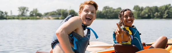 Giovane ed eccitato uomo rossa con la bocca aperta guardando la fotocamera vicino gioia donna afroamericana in giubbotto di salvataggio che punta con il dito durante la navigazione in kayak il giorno d'estate, banner — Foto stock