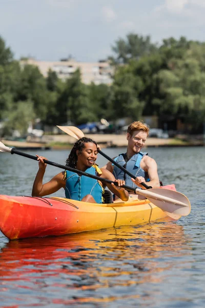Pareja interracial sin preocupaciones en chalecos salvavidas sosteniendo paletas y mirando el agua clara mientras navega en kayak deportivo durante el fin de semana de recreación en el día de verano - foto de stock