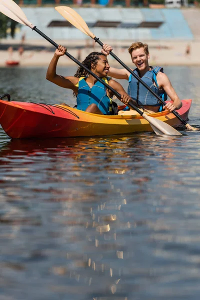 Charmante femme afro-américaine en gilet de sauvetage regardant homme rousse excité tout en naviguant en kayak sportif avec des pagaies le jour d'été sur le premier plan flou — Photo de stock