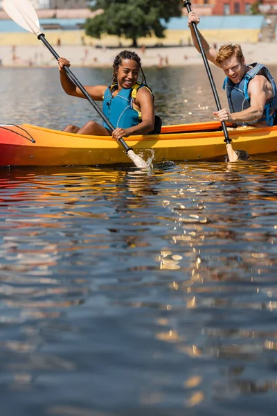 Felice donna africana americana e giovane uomo rossa in giubbotti di salvataggio guardando spruzzi d'acqua mentre pagaia in kayak sportivo durante il fine settimana estivo — Foto stock