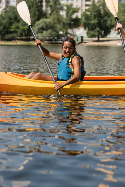 Вражена і весела афроамериканська жінка в житті жилет тримає весло під час плавання на каяку під час відпочинку у вихідні на міському озері в літній день — стокове фото