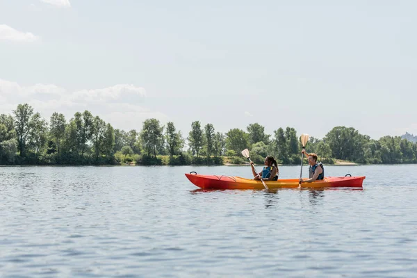 Vista lateral de la mujer afroamericana con el hombre joven en chalecos salvavidas remando en kayak deportivo a lo largo de la orilla del río verde durante la actividad recreativa en verano - foto de stock