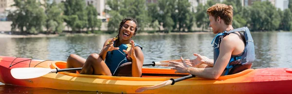 Alegre mujer afroamericana en chaleco salvavidas gesto cerca de joven pelirrojo mientras pasa el fin de semana de verano en el río y sentado en kayak deportivo, bandera - foto de stock
