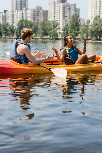 Femme afro-américaine impressionnée dans la vie gilet assis en kayak sportif près de jeune ami rousse et montrant wow geste sur le lac avec des bâtiments de la ville flous sur le rivage — Photo de stock