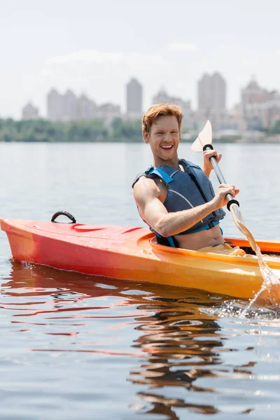 Young and overjoyed redhead man in life vest looking at camera and holding paddle while sailing in sportive kayak on lake with cityscape on blurred background — Stock Photo