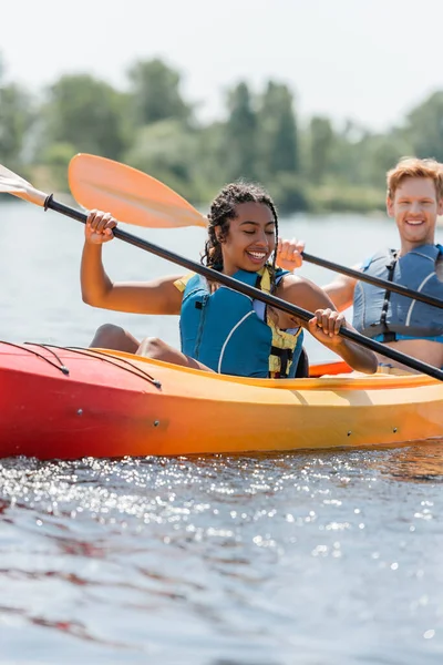 Femme afro-américaine insouciante et charmante dans le gilet de sauvetage tenant la pagaie et la voile en kayak sportif près du jeune homme rousse souriant sur fond flou — Photo de stock