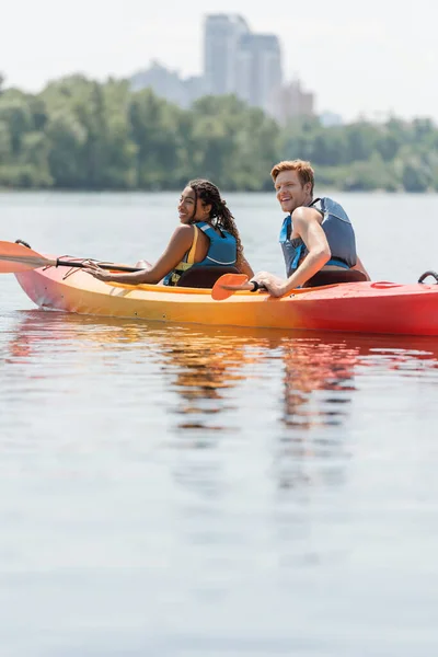 Felice donna africana americana e giovane, sorridente uomo rossa in giubbotti di salvataggio seduto in kayak vicino alle pagaie e guardando lontano durante il fine settimana estivo sul fiume calmo — Foto stock