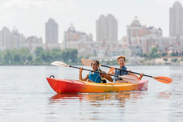 Happy and impressed multiethnic friends in life vests holding paddles and smiling at camera in sportive kayak on river with scenic cityscape on blurred background — Stock Photo
