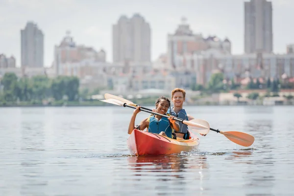 Despreocupados pareja interracial en chalecos salvavidas sosteniendo paletas y mirando a la cámara mientras navega en kayak deportivo en el lago con paisaje urbano borroso en el fondo - foto de stock