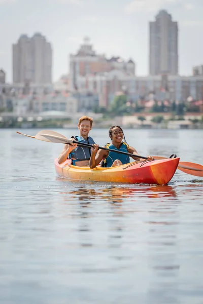 Carefree and enchanting african american woman and young redhead man in life vests holding paddles and sailing in sportive kayak on river with scenic cityscape on blurred background — Stock Photo