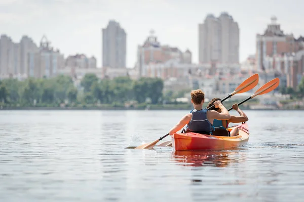 Vista trasera de la pareja multiétnica activa en chalecos salvavidas remando en kayak deportivo en la superficie del agua tranquila en el día de verano con vista a edificios urbanos borrosos a orillas del río - foto de stock