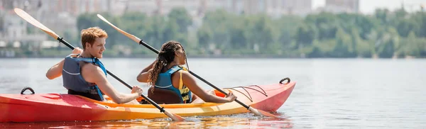 Hombre joven y pelirrojo y mujer afroamericana en chalecos salvavidas navegando en kayak deportivo con paletas en río tranquilo durante la recreación acuática en el día de verano, bandera - foto de stock