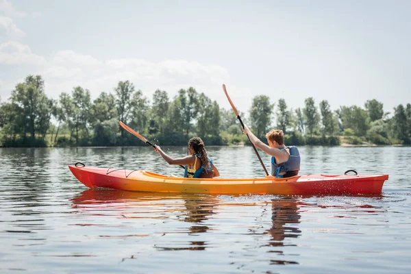 Vista lateral de pareja multiétnica activa en chalecos salvavidas sosteniendo paletas mientras navega en kayak deportivo en el río con pintoresca orilla con árboles verdes en verano - foto de stock