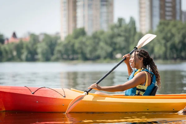 Vista laterale della donna afroamericana attiva e affascinante in giubbotto di salvataggio vela in kayak sportivo con pagaia il giorno del fine settimana estivo su sfondo sfocato — Foto stock