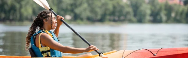 Вид збоку активної і щасливої афроамериканської жінки в житті жилет тримає весло і плаває в каяку, проводячи літні вихідні на мальовничій річці, банер — стокове фото