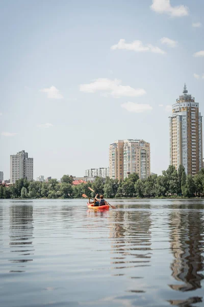 Vista trasera de la pareja deportiva interracial en chalecos salvavidas navegando en kayak a orillas del río con árboles verdes y edificios contemporáneos durante la actividad recreativa en el fin de semana de verano - foto de stock