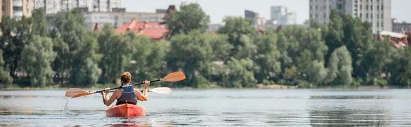 Vue arrière des amis multiethniques actifs dans les gilets de sauvetage naviguant en kayak sportif avec pagaies sur la rivière de la ville avec rive verte pendant le week-end de loisirs en été, bannière — Photo de stock