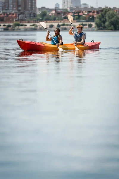 Coppia multirazziale positiva e attiva in giubbotti di salvataggio che tengono pagaie durante la navigazione in kayak sportivo sulla superficie dell'acqua calma durante il fine settimana ricreativo estivo sul lago cittadino — Foto stock