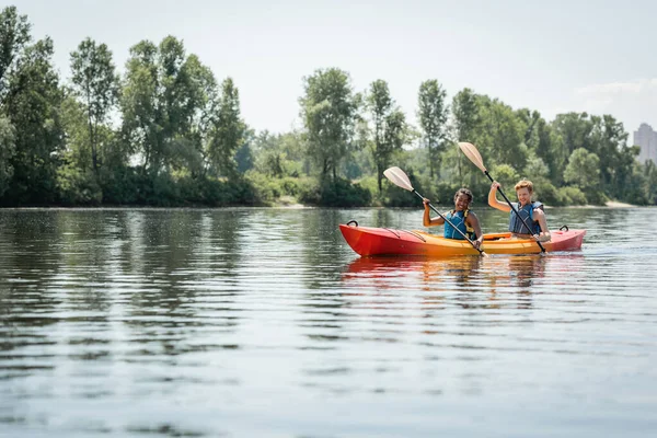 Active and happy multiethnic couple in life vests holding paddles while sailing in sportive kayak on lake with green trees on scenic shore during water recreation in summer — Stock Photo